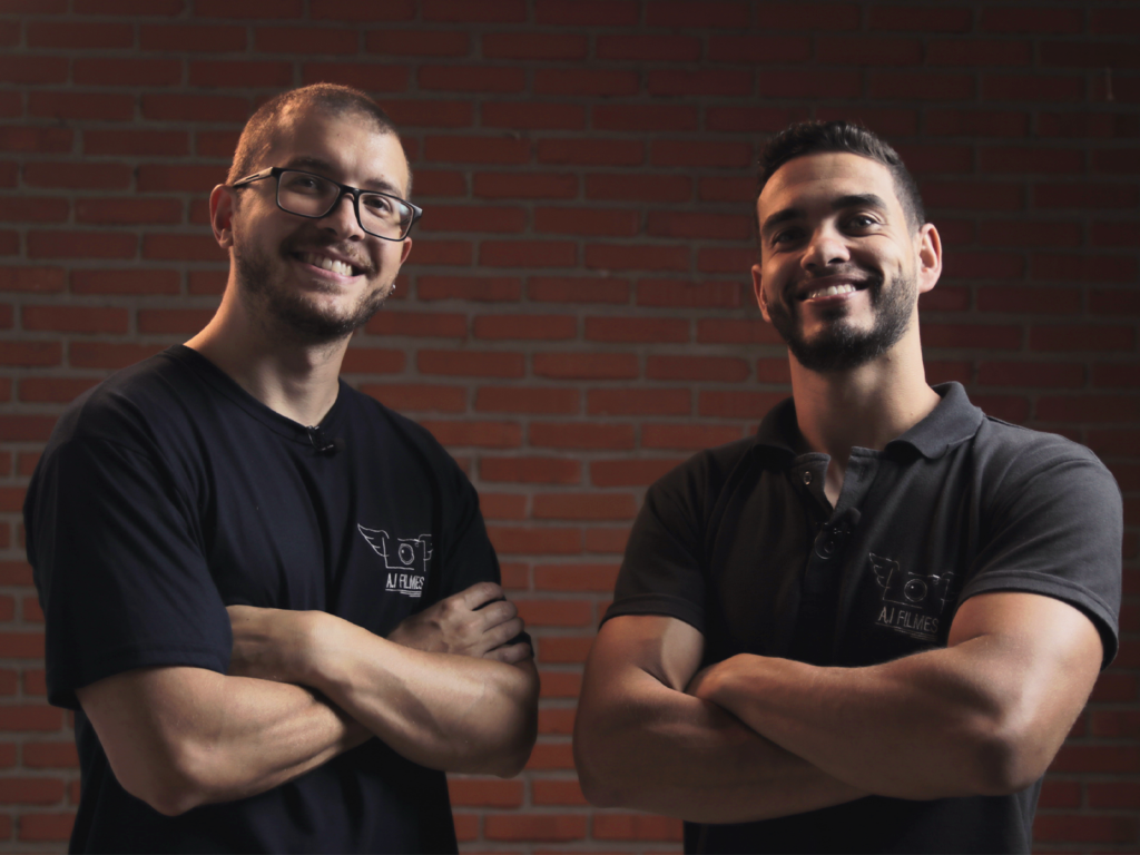 Dois homens de camisa preta, sorrindo, de braços cruzados olhando para a lente